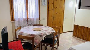 A dining area with a round table covered by a floral tablecloth, wooden chairs, a red rug, and a television on a stand.
