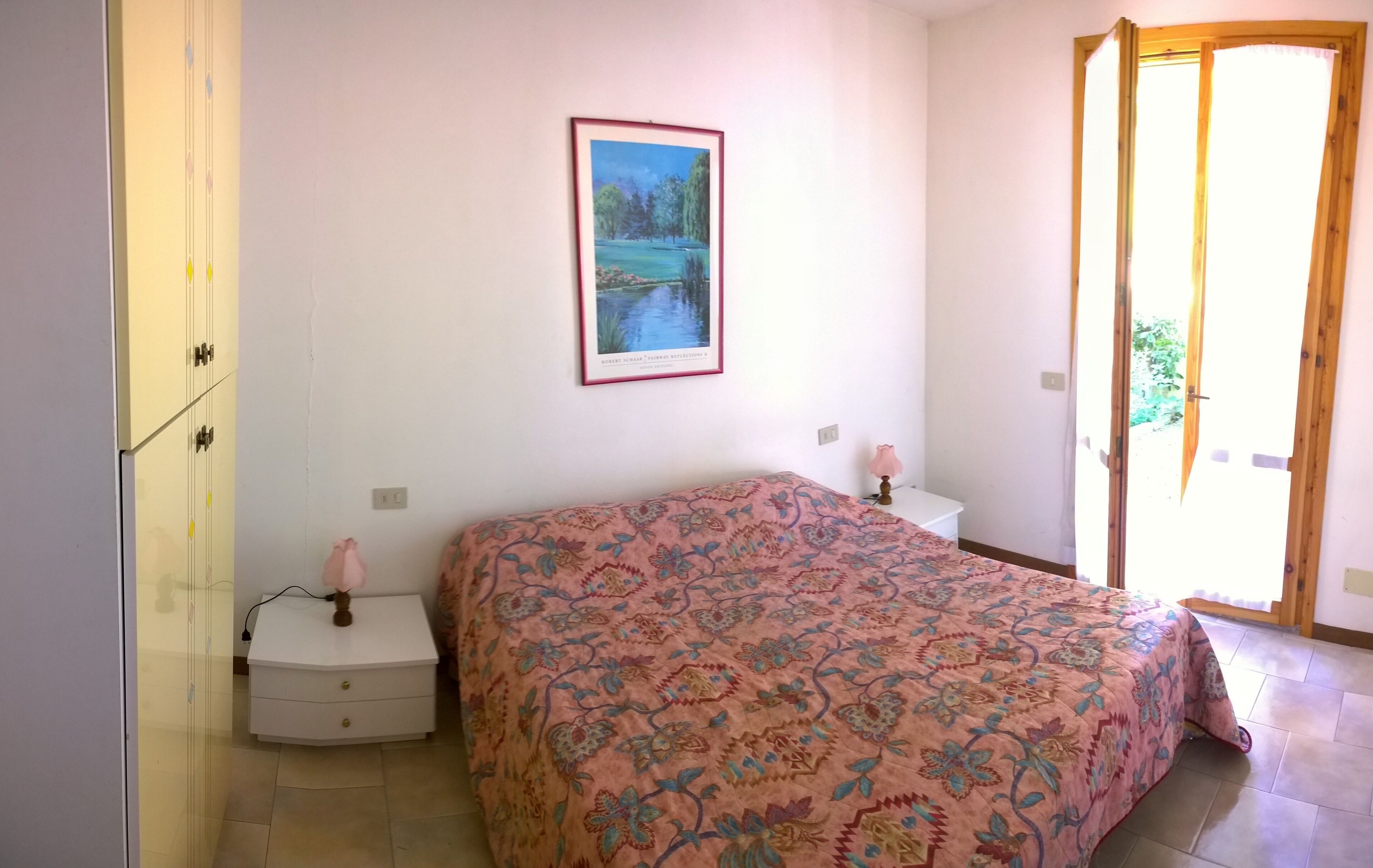 Un dormitorio con cama, mesita de noche, lámpara, un cuadro en la pared y una puerta que da a un espacio al aire libre.