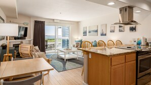 A modern kitchen with a central island, wooden cabinets, and a built-in oven. There is a dining area with a table and chairs, and a large window offering a view of the outdoors. - Stunning views from this bayfront condo in the Taft district of Lincoln City! (Lincoln City)