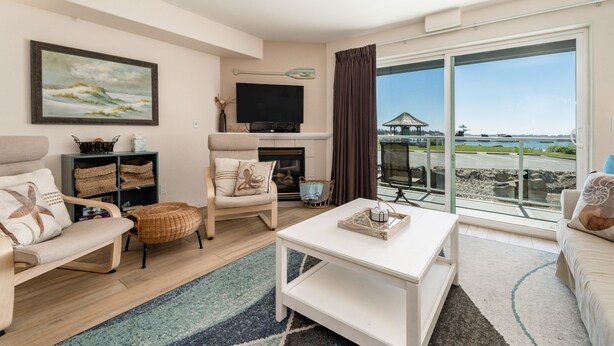 A living room with a sofa, armchair, and coffee table, a fireplace, a television, and a large window with a view of the beach and a gazebo. - Stunning views from this bayfront condo in the Taft district of Lincoln City! (Lincoln City)