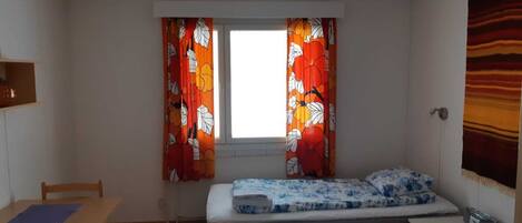 A small, simple room with two beds, a desk, and a window with curtains.