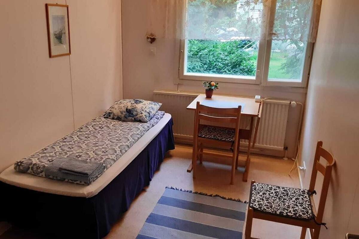 A small room with a single bed, a chair, a table, and a window with a view of greenery.
