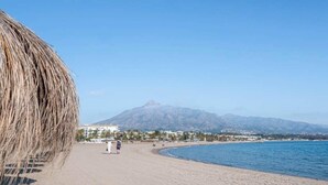 Ein Strand mit einem Berg im Hintergrund, einer reetgedeckten Struktur links und einigen Personen, die spazieren gehen.