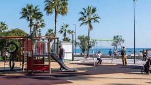 Ein Spielplatz mit Rutschen und Schaukeln, Palmen und freiem Blick auf das Meer.