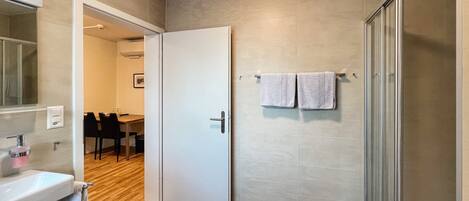 A modern bathroom with a shower, a sink, and a toilet. There is a door leading to a dining area.