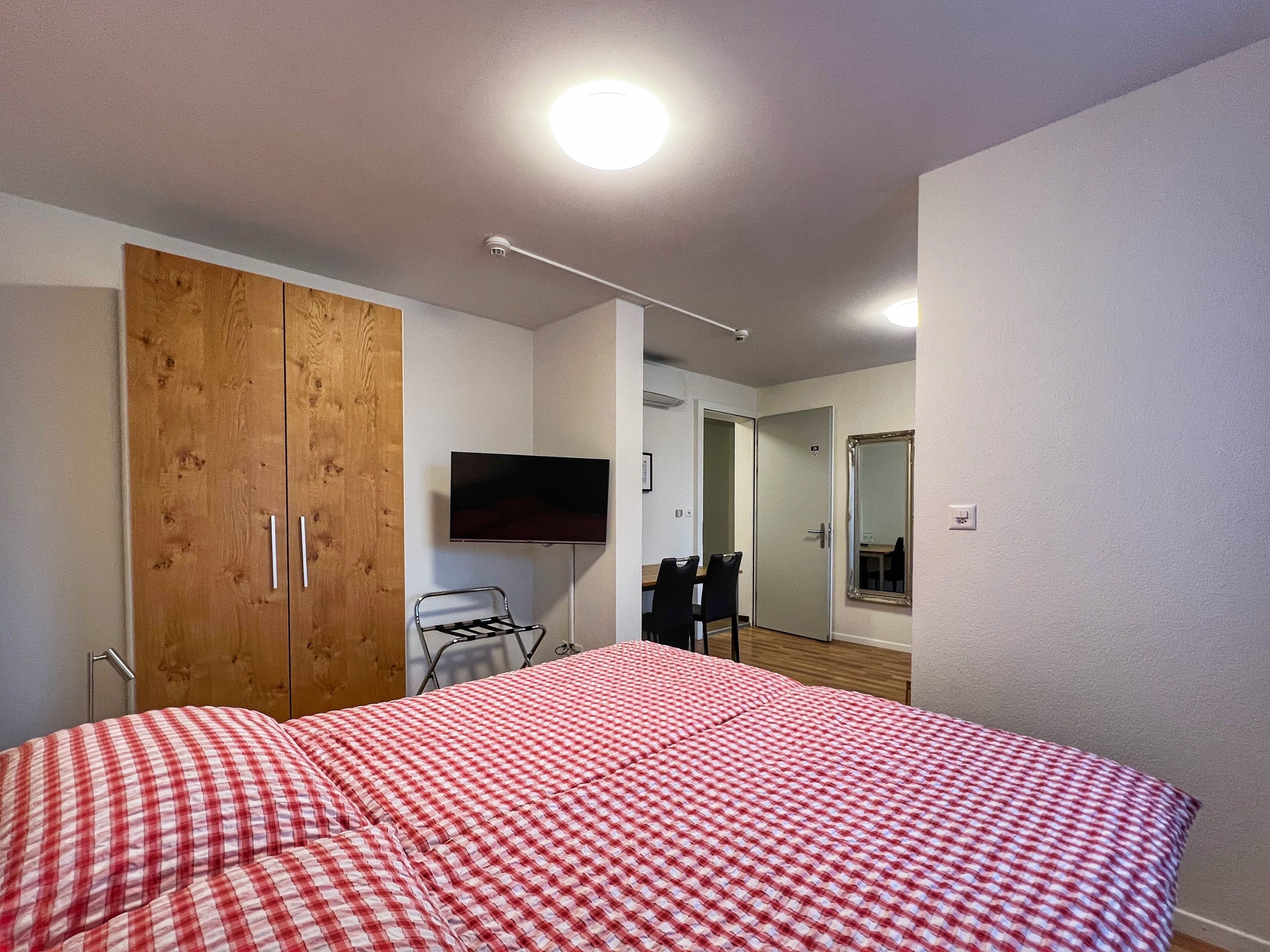 A bedroom with a checkered bedspread, a wooden wardrobe, a television, and a mirror.