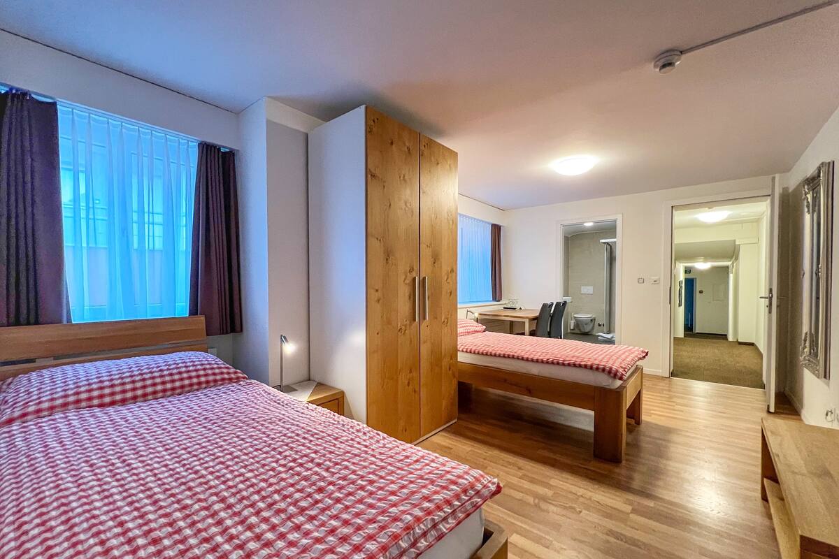 A bedroom with two beds, a wooden wardrobe, a desk, and a bathroom visible through an open door.
