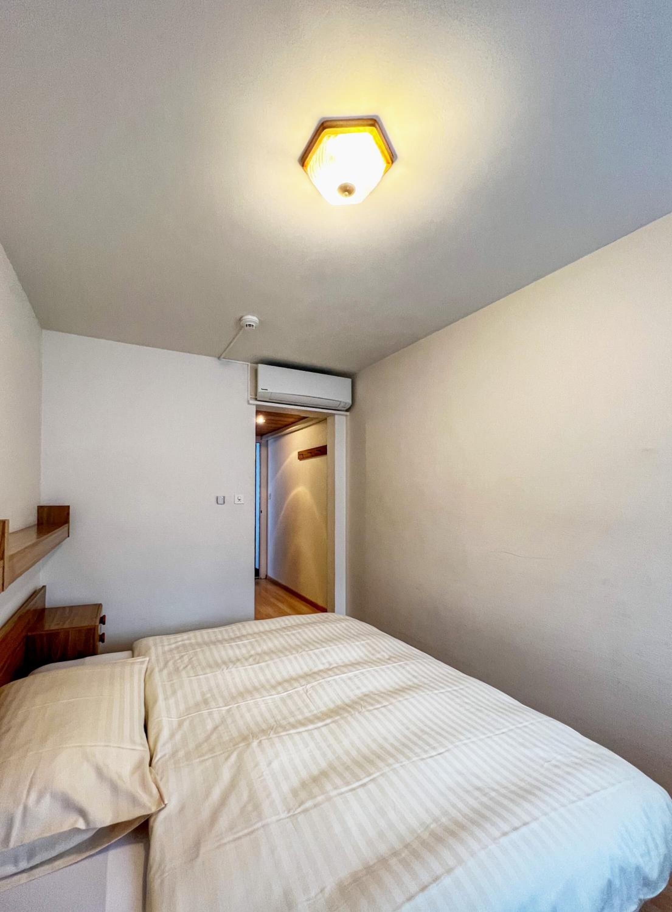 A small, sparsely furnished room with a bed, a wooden nightstand, a wall-mounted air conditioner, and a hallway leading to another room.