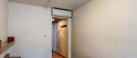 Una habitación pequeña y poco amueblada, con una cama, una mesita de noche de madera, aire acondicionado instalado en la pared y un pasillo que conduce a otra habitación.