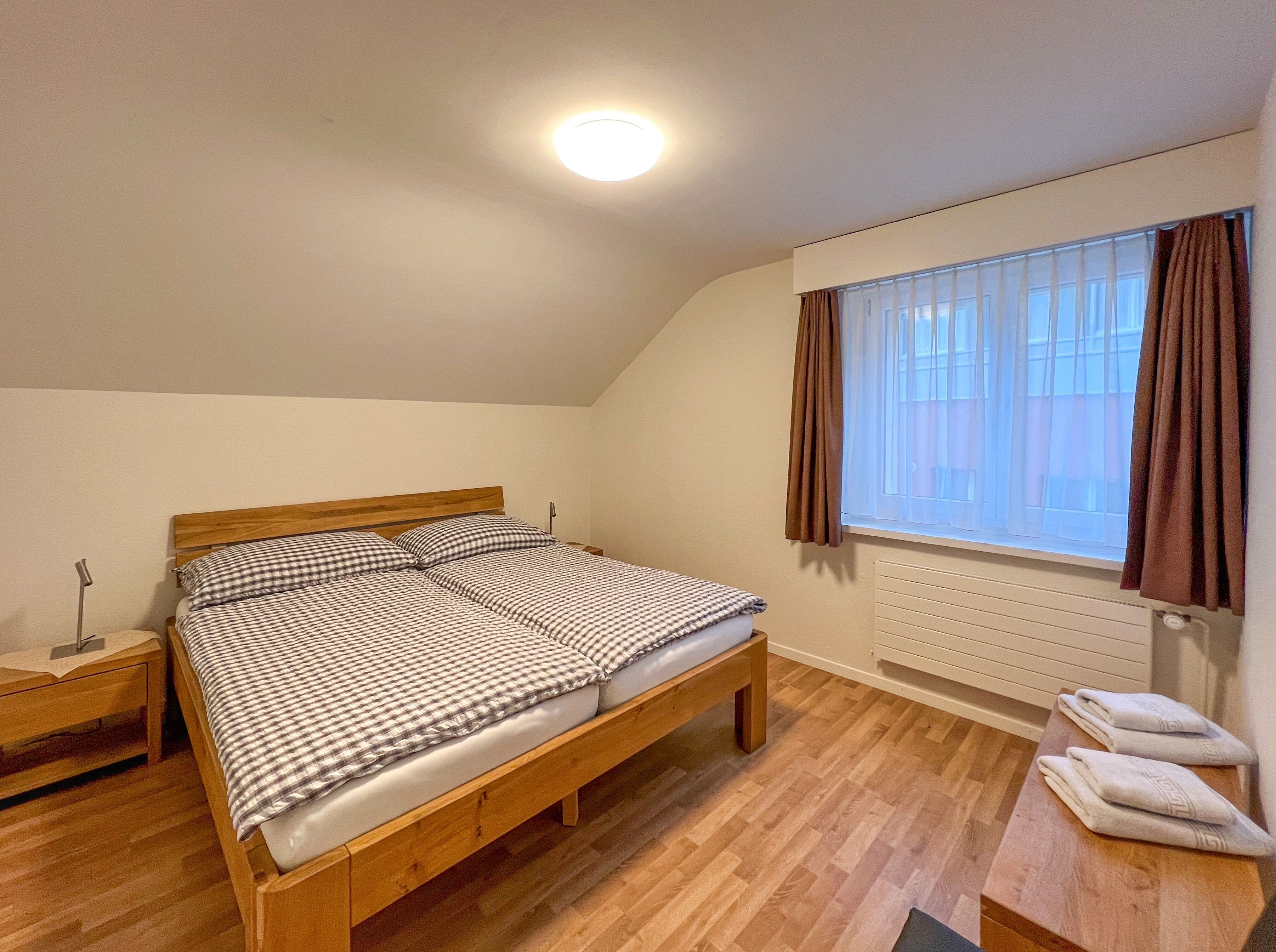 A bedroom with a wooden bed, a nightstand, a window with curtains, and a radiator.