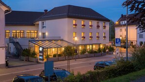 A hotel with a covered entrance, a parking sign, and a street view with cars and greenery. - Hotel Hecht (Rheineck)