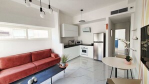 A modern kitchen with white cabinets, a stainless steel refrigerator, and a built-in oven. There is a red sofa and a small table with a plant. - Design 2 Bdr Apartment - Habima TL60 (Tel Aviv)