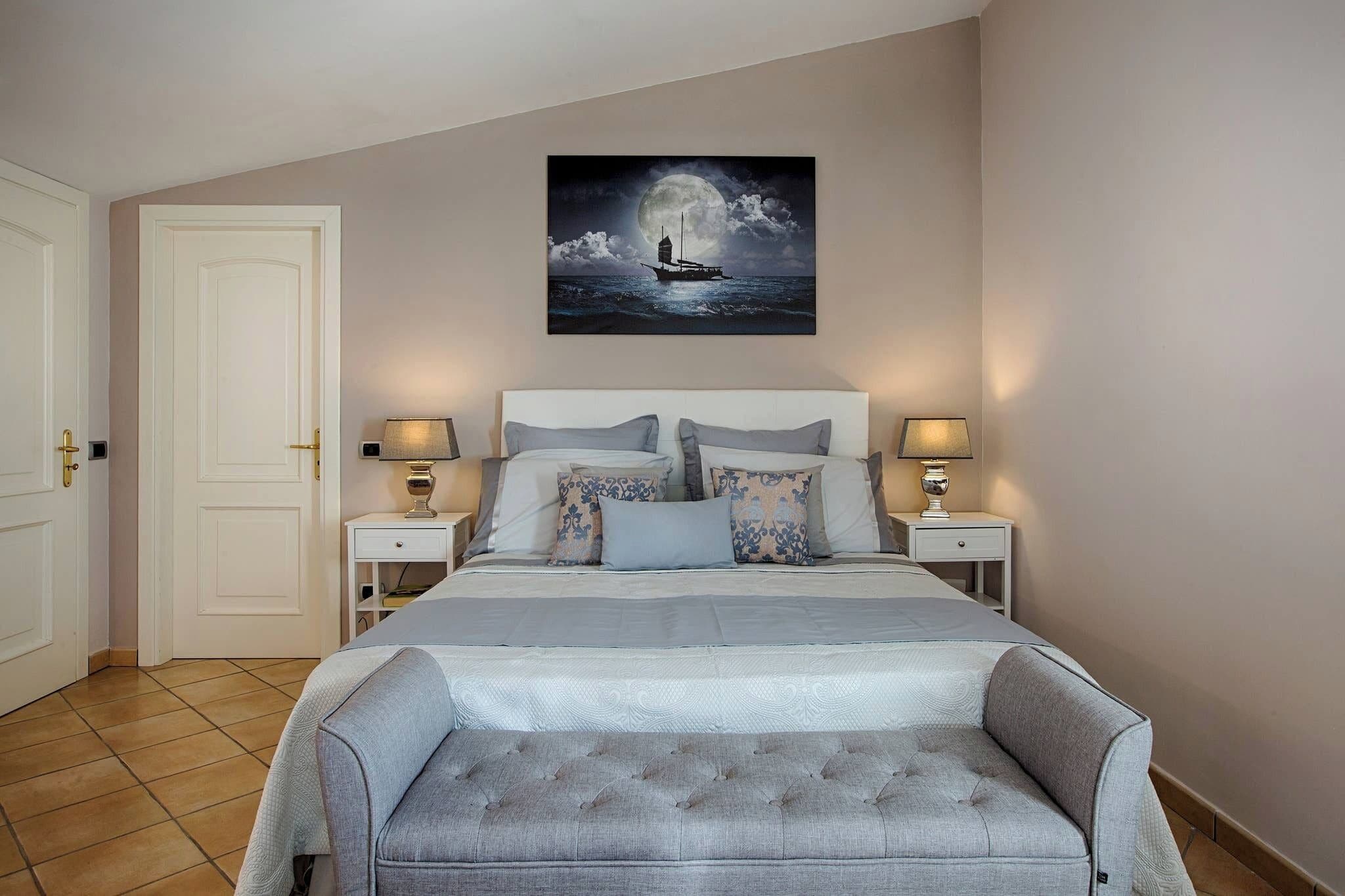Un dormitorio con una cama, dos mesitas de noche, un banco, una puerta y una obra de arte colgada en la pared.
