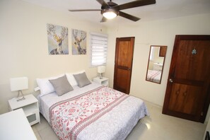 A bedroom with a bed, nightstands, a ceiling fan, a wooden door, and two wall-mounted paintings. - Hostal Caribean Apart (Santo Domingo)