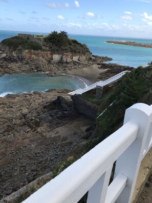 Paisagem costeira com terreno rochoso, uma ponte branca e um farol ao longe.