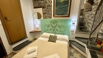 Un dormitorio con pared de piedra, cama con ropa de cama blanca, cabecera verde y una pintura de paisaje marino.