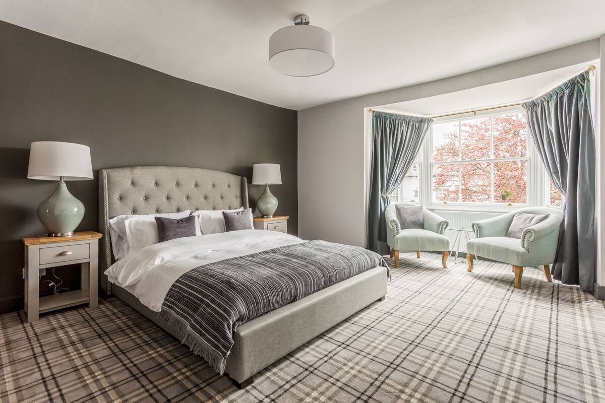 A bedroom with a tufted headboard bed, two bedside tables with lamps, a plaid-patterned floor, a window with curtains, and a chair by the window.