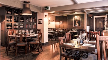 Um restaurante com mesas e cadeiras de madeira, garrafas de vinho nas prateleiras e um relógio na parede.