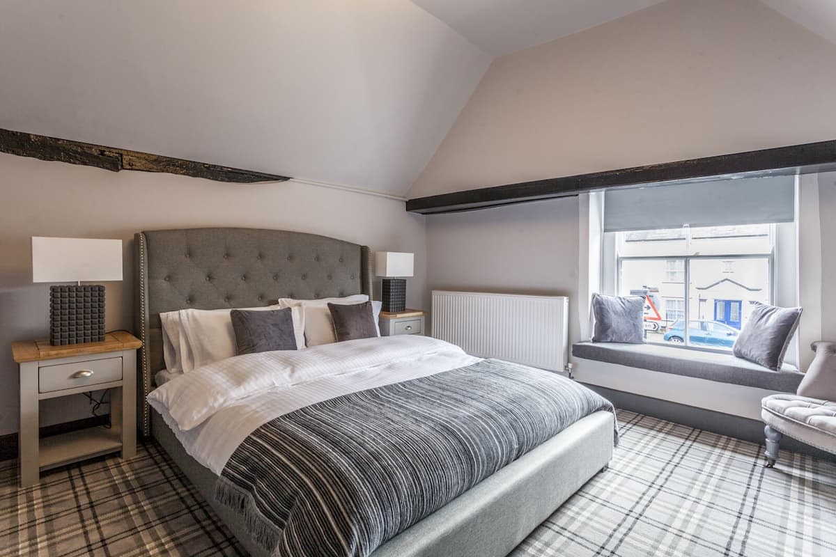 A bedroom with a large bed, a nightstand, a chair, a window with a view of the street, and a radiator.