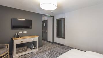 A hotel room with a bed, a TV mounted on the wall, a wooden bedside table, a small console table, a chair, and a door leading to another room.