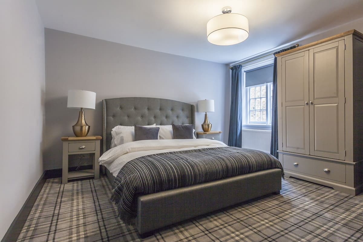 A bedroom with a tufted headboard, a large bed with a striped bedspread, two bedside tables with lamps, a wardrobe, and a window with curtains.