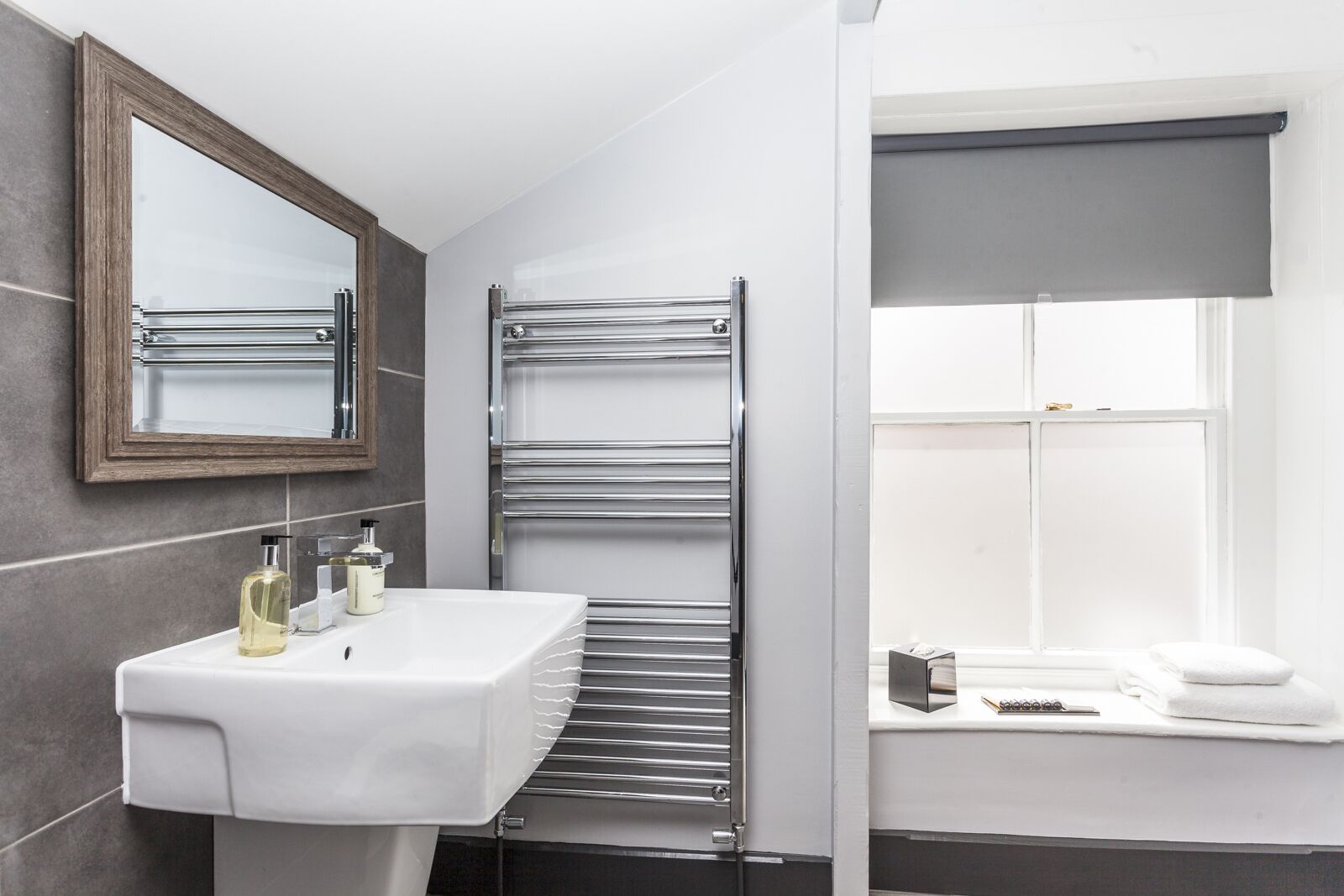 A modern bathroom with a white sink, a chrome towel rack, a mirror, and a window with a gray blind.