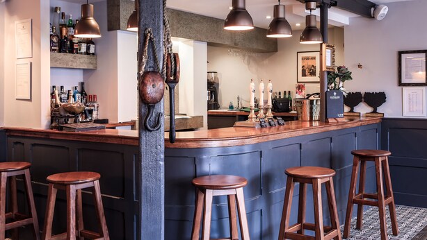 Zona de bar amb tamborets de fusta, il·luminació penjant de coure i una varietat de botelles i vasos a les estanteries.