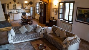 A cozy living room with two patterned sofas, a wooden coffee table, and a stone fireplace. A kitchen is visible in the background with white cabinets and a refrigerator. - Podere Le Rote Private Farmhouse (Castelnuovo di Val di Cecina)