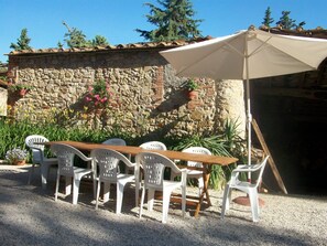 An outdoor patio with a wooden table and white plastic chairs, a beige umbrella, and a stone wall with potted plants.