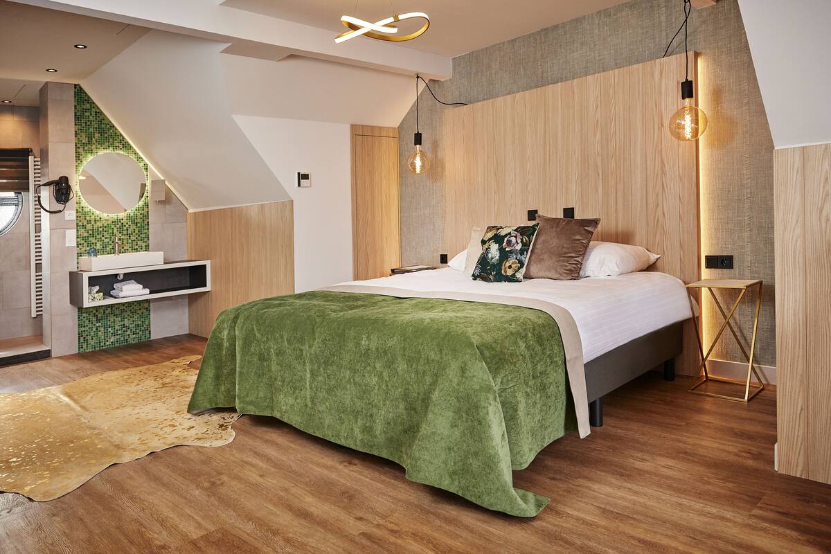 Un dormitorio moderno con una cama, suelo de madera, un plaid verde, un espejo redondo y un estante pequeño.