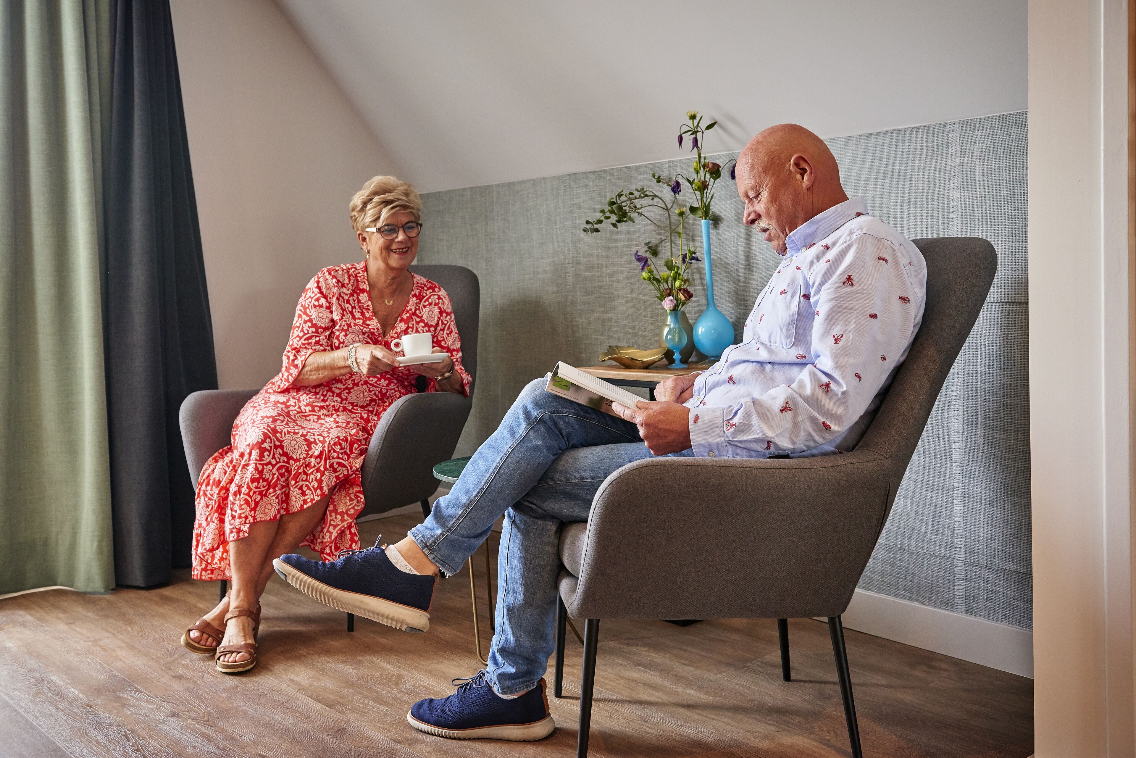 Twee personen zitten in een kamer met een houten vloer, een grijze fauteuil en een kleine tafel met een bloevas.