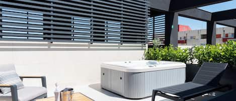 A modern outdoor seating area with a hot tub, lounge chairs, and a wooden table with a drink container.