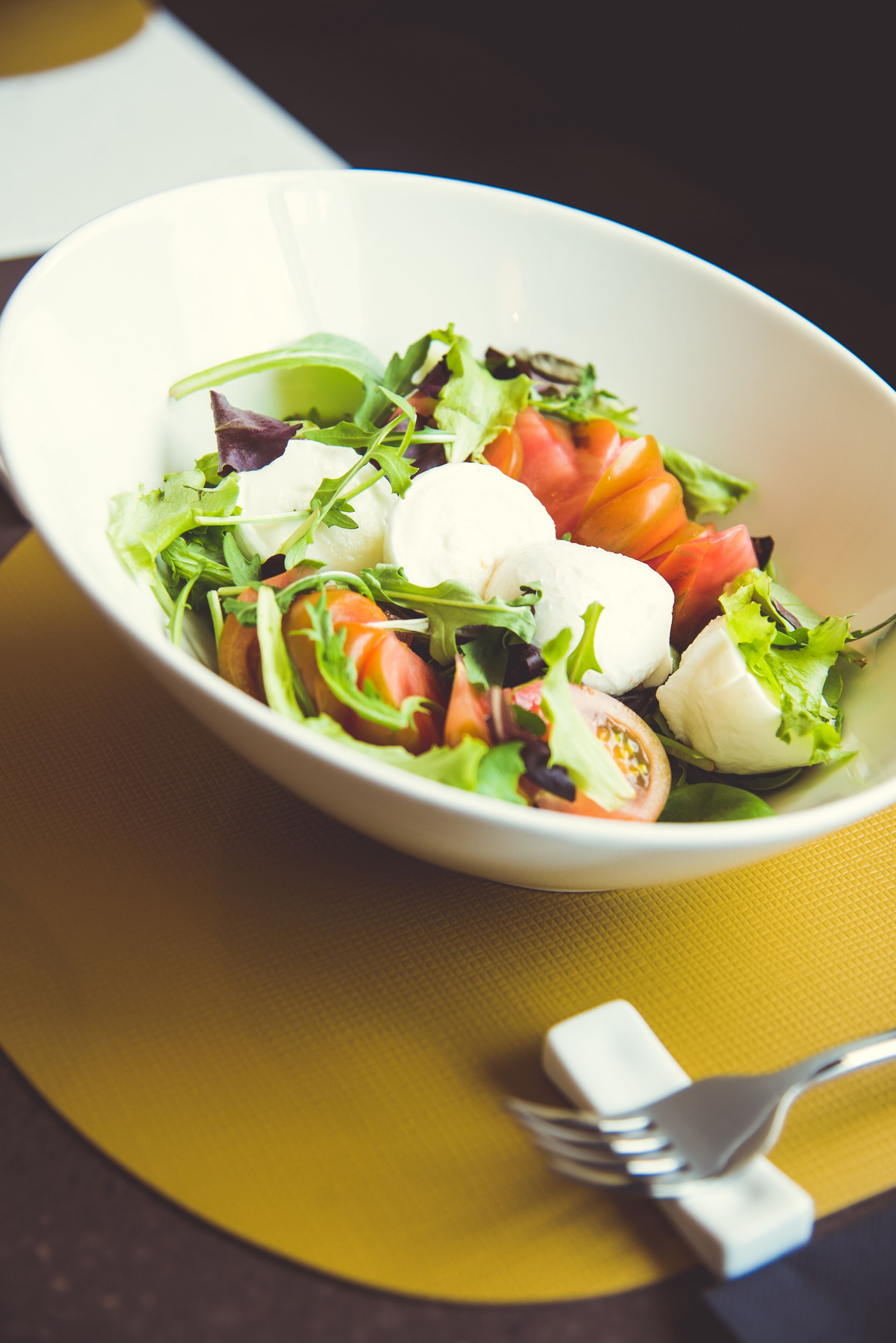 A white bowl of salad with various greens, tomatoes, and mozzarella on a yellow napkin.