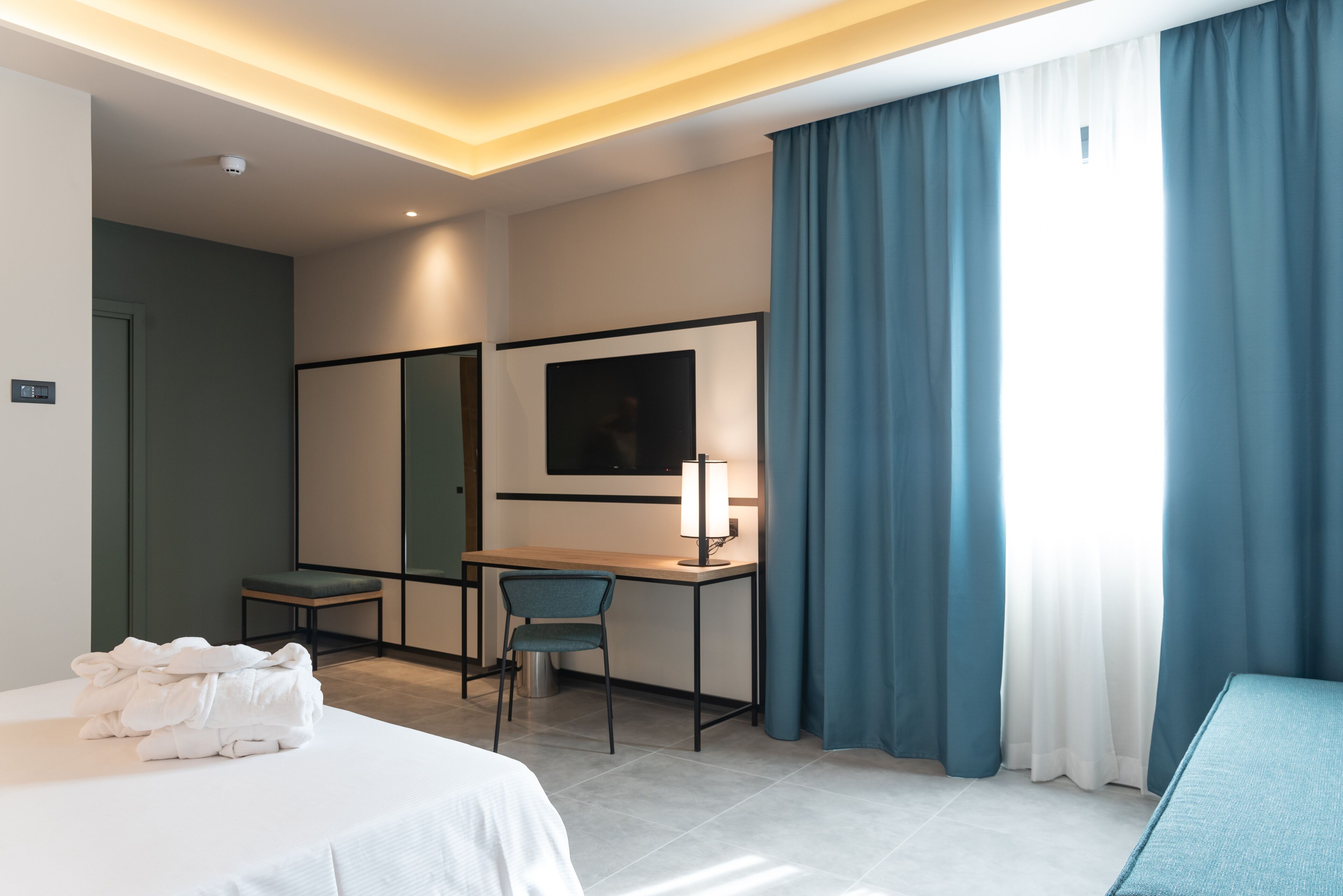 A hotel room with a bed, a desk with a chair, a TV, and a window with curtains.