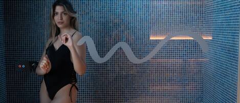 A person in a black swimsuit standing in front of a tiled wall with a wavy pattern.