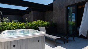Un espace moderne aménagé à l’extérieur avec un jacuzzi, une chaise longue et un banc, le tout entouré de verdure et adossé à la façade contemporaine d’un bâtiment.