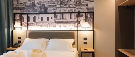 Quarto de hotel com uma cama grande, duas luminárias de cabeceira, mesa de cabeceira com telefone e uma obra de arte na parede retratando uma paisagem urbana.