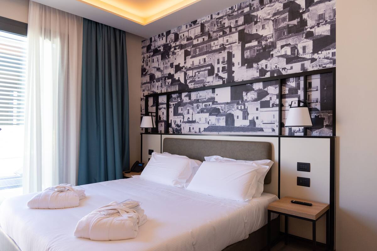 A hotel room with a large bed, two bedside tables, a nightstand, a wall-mounted lamp, and a window with curtains.