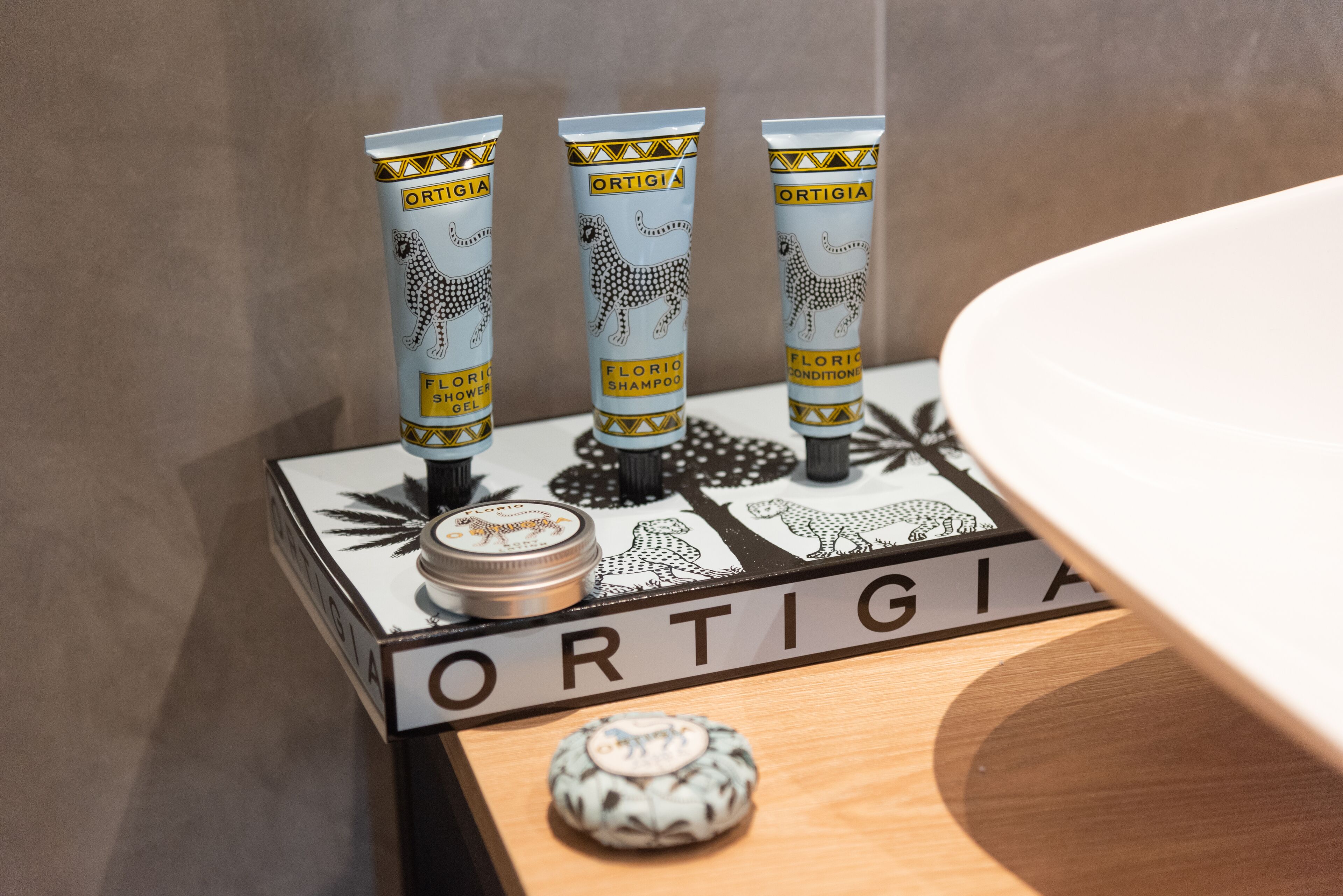 Three tubes of 'ORTIGIA' shower gel and conditioner on a wooden shelf with a nameplate.