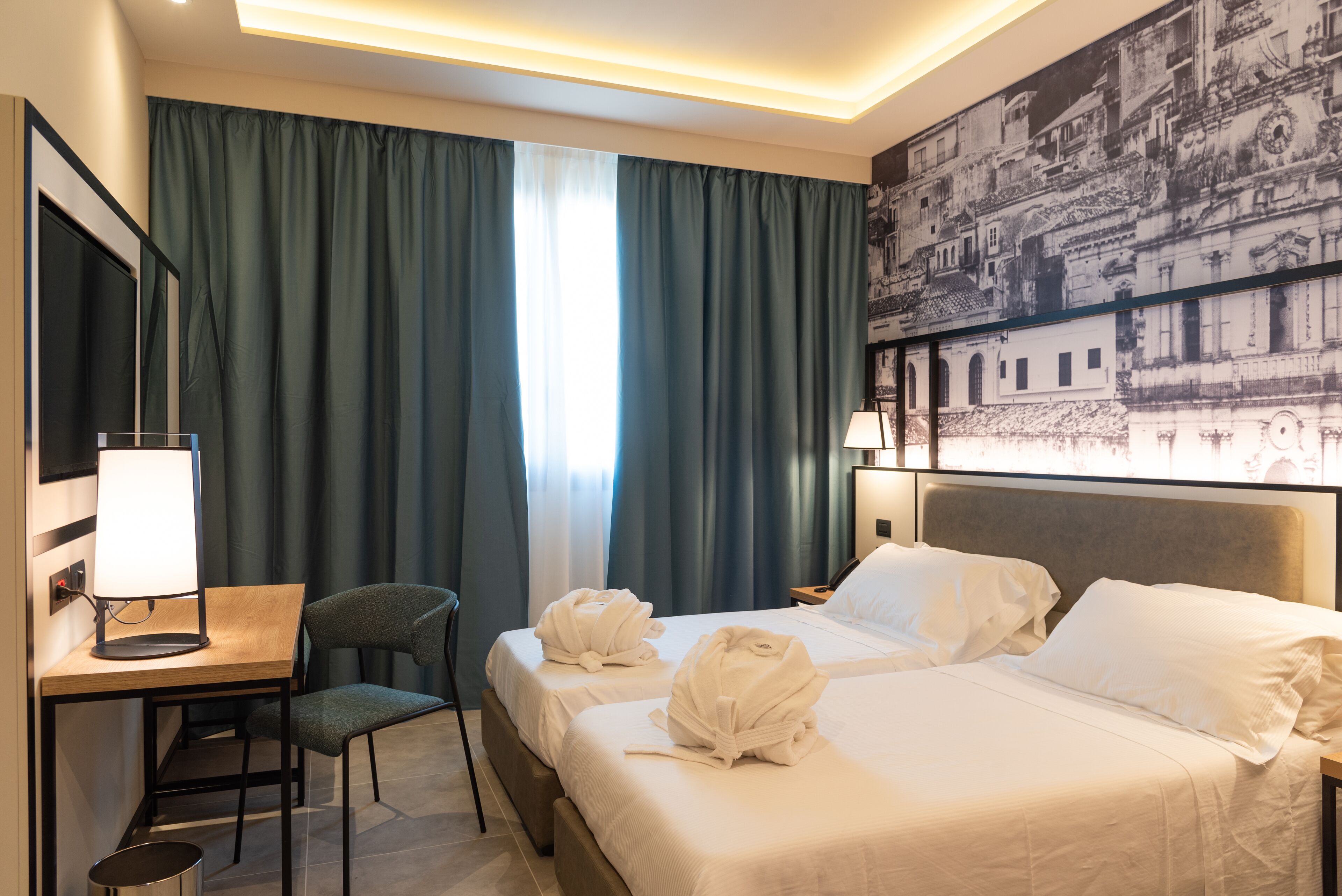 Une chambre d’hôtel avec deux lits, un bureau avec une lampe, une chaise et un grand tableau représentant un paysage urbain accroché au mur.