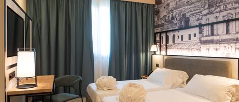 Habitación de hotel con dos camas, un escritorio con lámpara, una silla y un cuadro grande con un paisaje urbano en la pared.