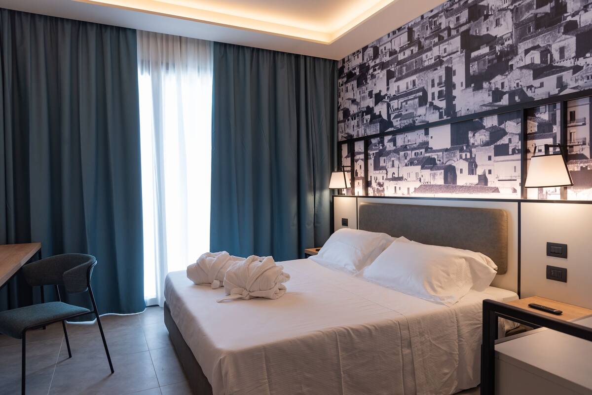 A hotel room with a neatly made bed, a desk with a chair, and a large window with curtains.