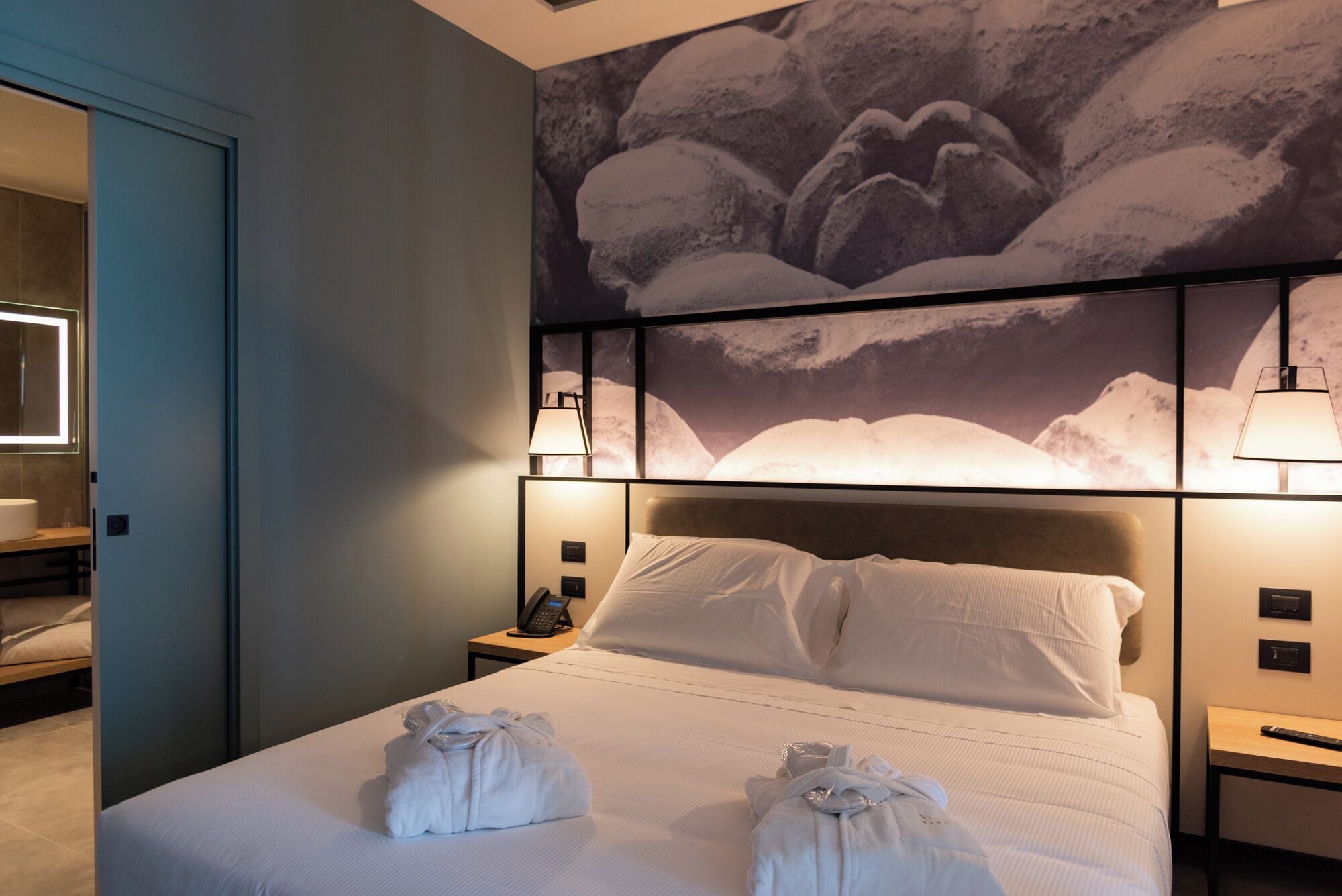 A hotel room with a large bed, a headboard with a mountain landscape mural, bedside lamps, and a nightstand with a phone and a remote control.