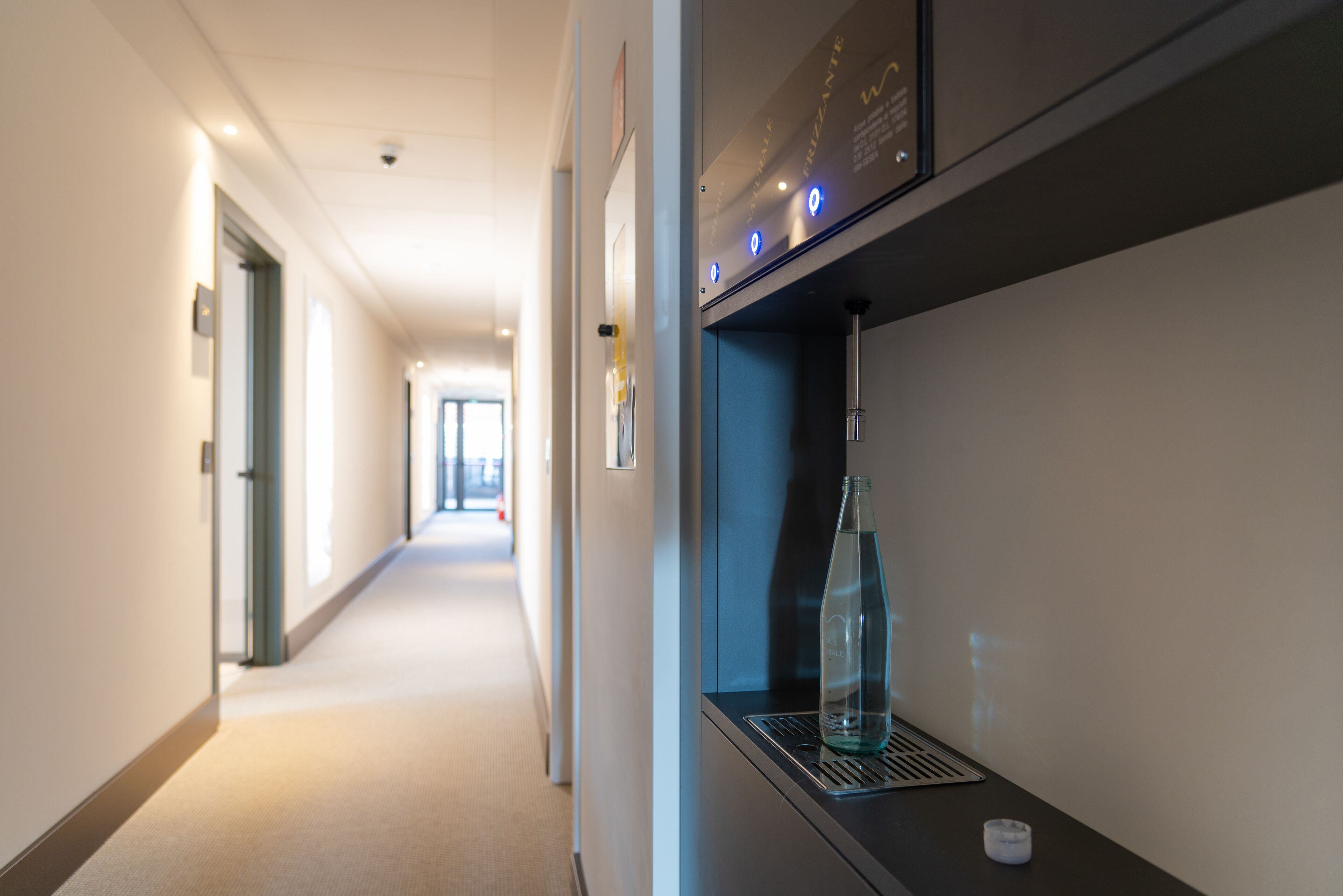 Un couloir d’hôtel avec un distributeur d’eau et une bouteille fixée au mur.