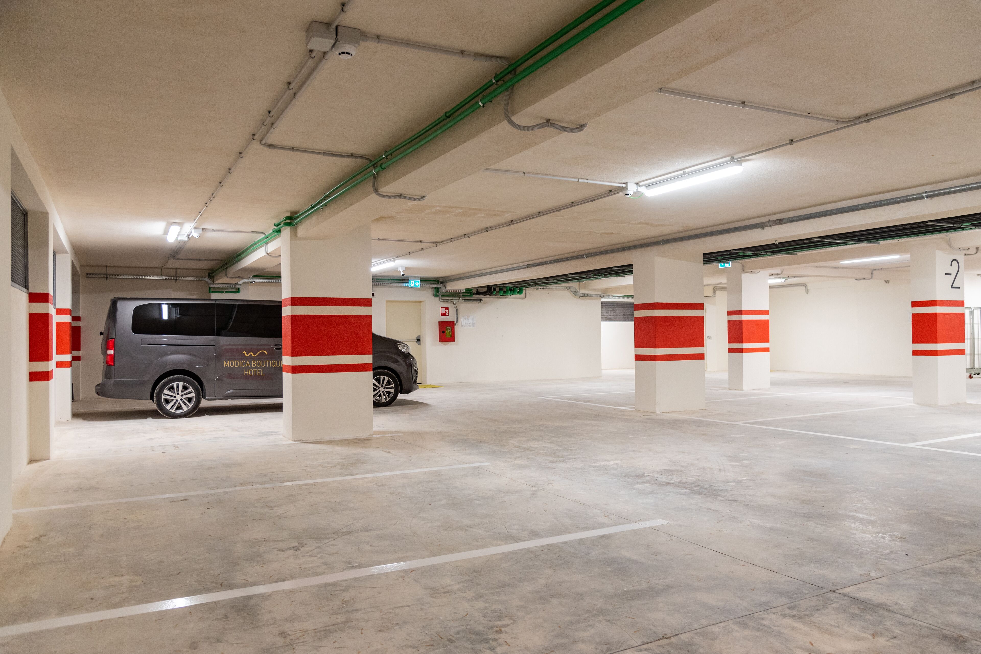 A parking garage with a grey van parked in a designated space, white pillars with red stripes, and clear ceiling pipes.