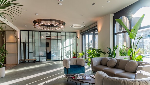 Un hall d’entrée moderne avec un fauteuil bleu, un canapé beige et une table basse sur laquelle sont posés des magazines. On y trouve également des plantes en pot et une grande fenêtre laissant entrer la lumière naturelle.
