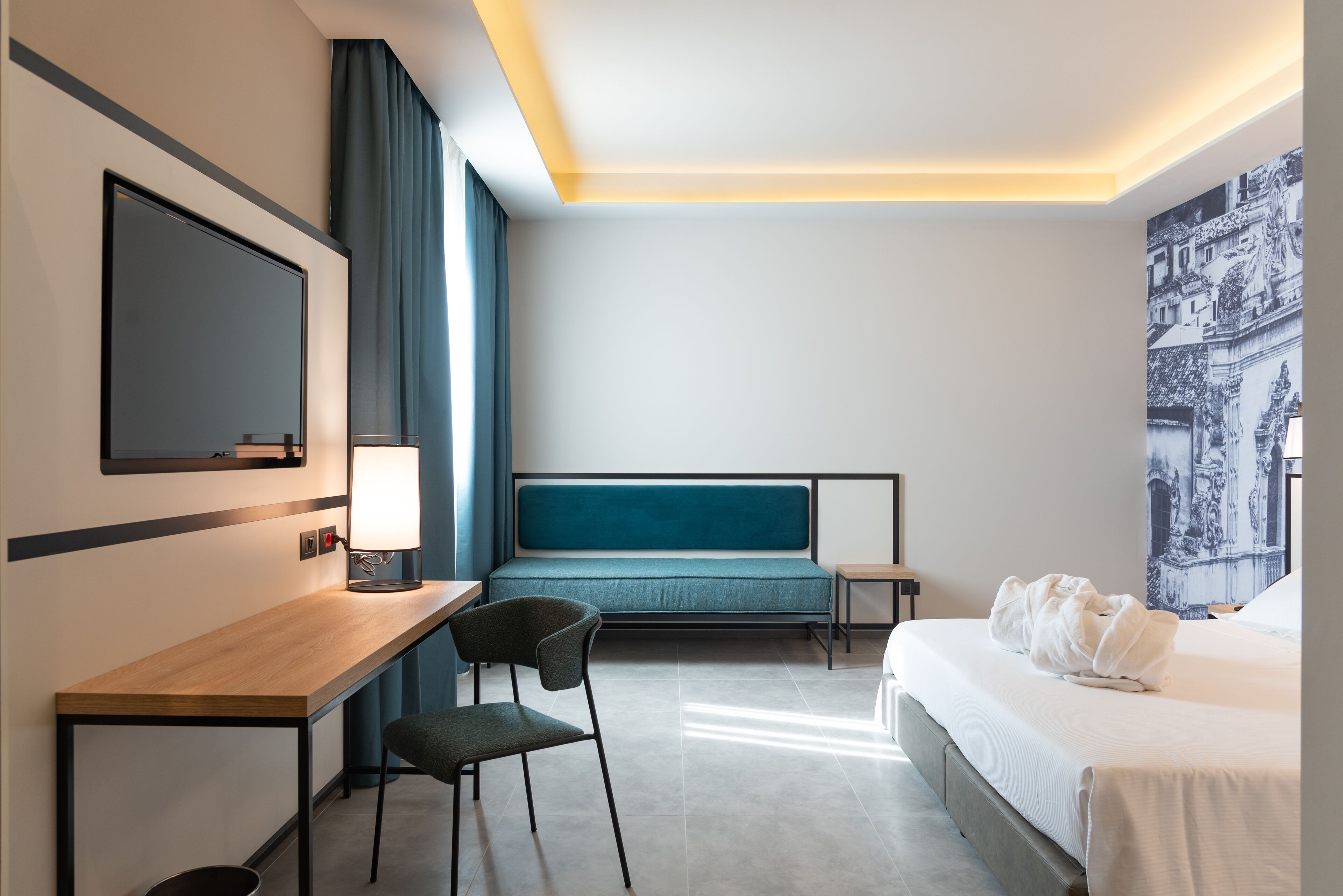 A hotel room with a bed, a desk with a chair, a TV, and a large window with curtains.