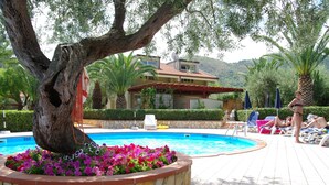A swimming pool surrounded by stone flower beds, palm trees, and a building with a red roof. - Relaxing Apartment for 2 People in Residence (Cefalù)
