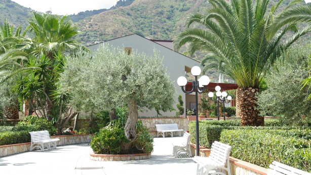 An outdoor courtyard with benches, palm trees, and a building with a stone facade. - Relaxing Apartment for 2 People in Residence (Cefalù)