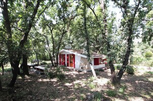 Una cabaña rústica rodeada de árboles frondosos, con un muro de piedra y detalles en rojo.